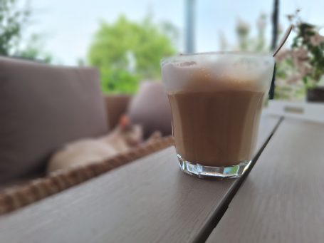 Ein Glas Kaffee steht auf einem Holztisch, im Hintergrund eine verschwommene Sitzecke.