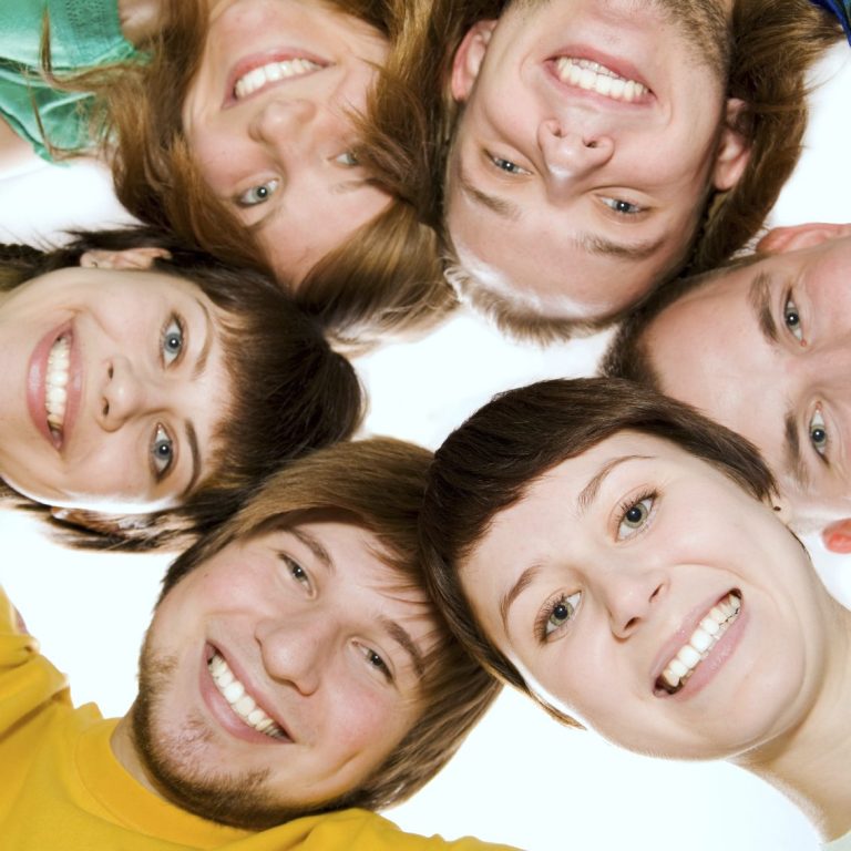 Sechs fröhlich lächelnde junge Menschen bilden einen Kreis, ihre Gesichter sind nah beieinander und sie blicken nach unten in die Kamera.